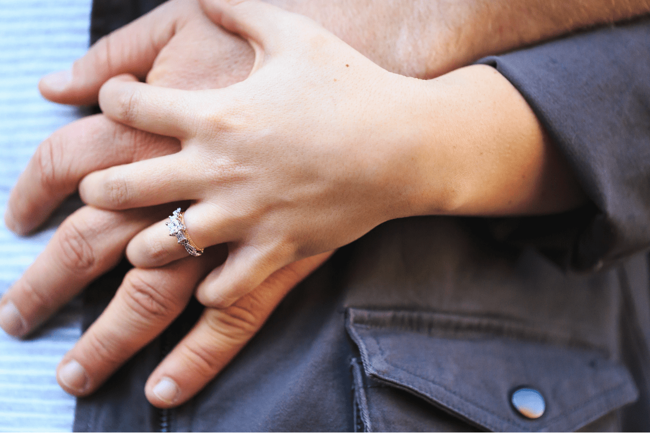 A woman’s hand with a three stone intertwining engagement ring is on top of her significant other’s hand