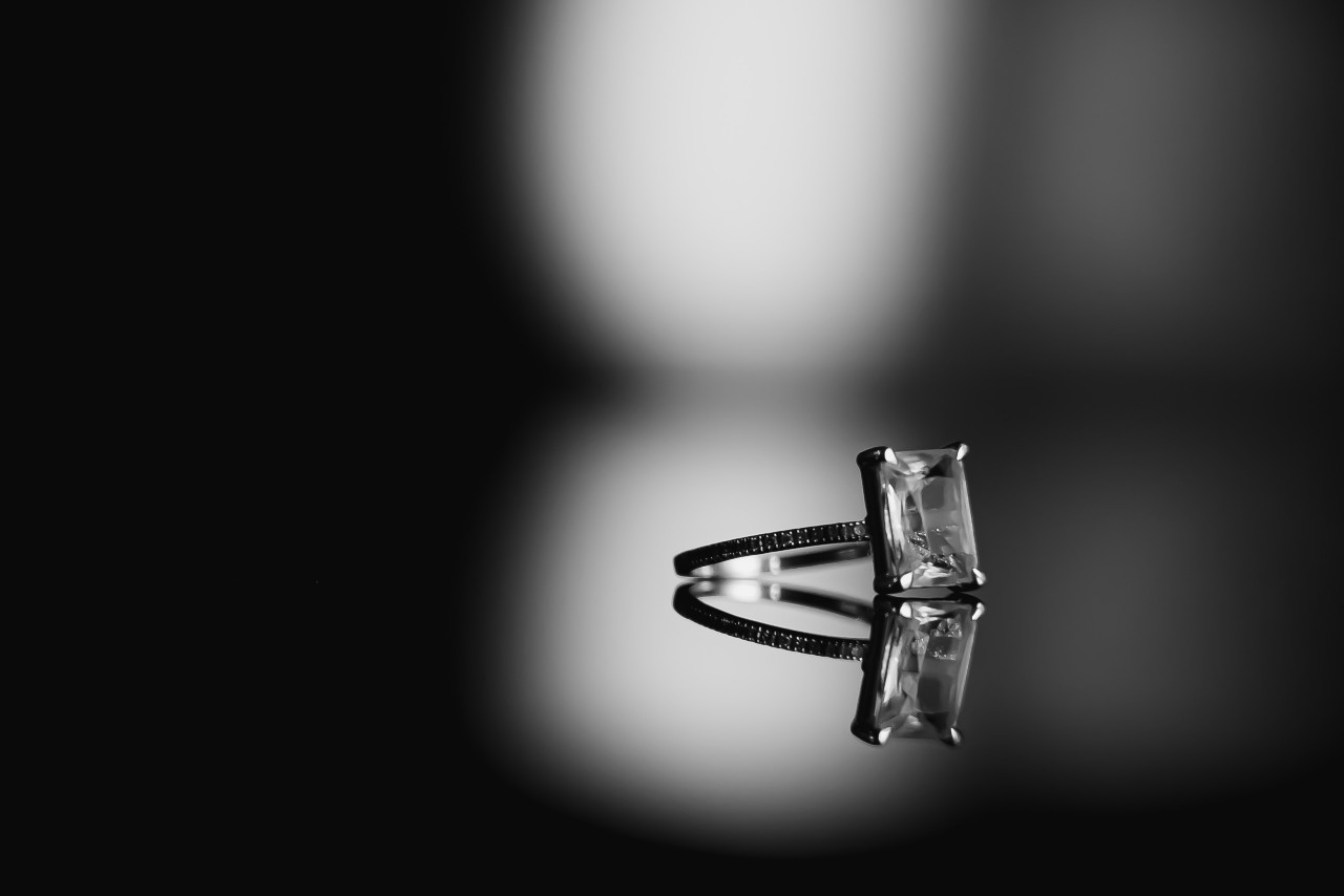 A radiant cut diamond solitaire engagement ring sits on a reflective black surface