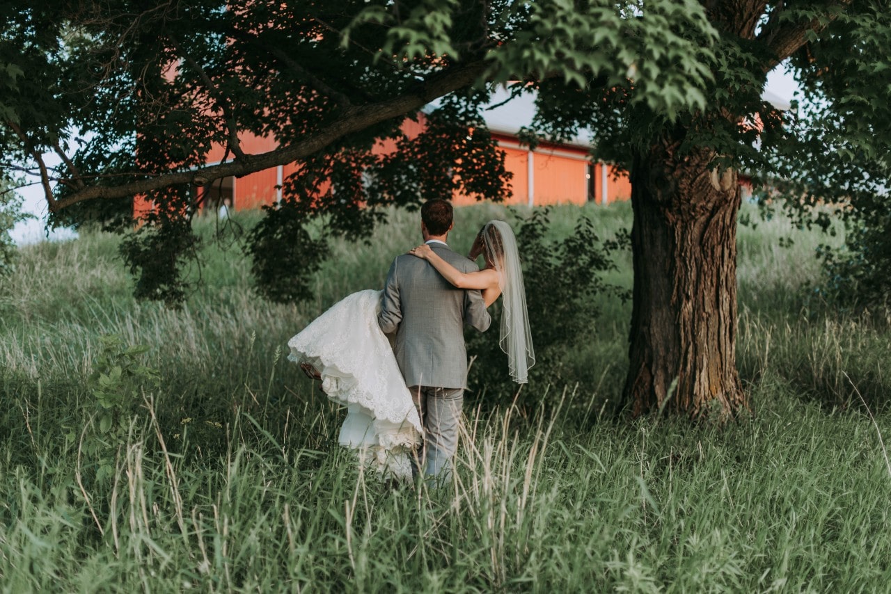 Groom holding a bride standing in tall grass under a tree with a barn in the background