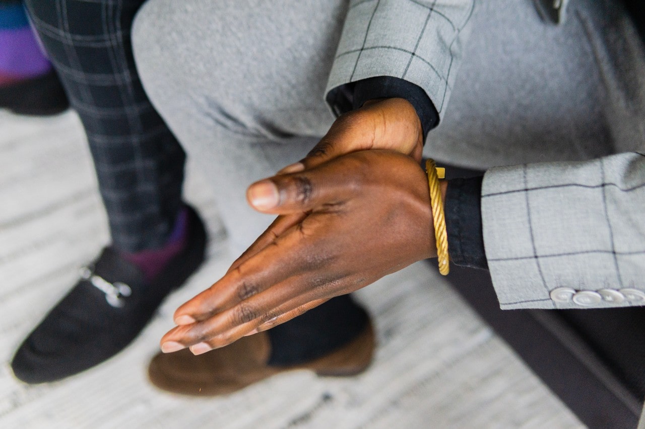 A man in a gray suit, pressing his hands against each other, a yellow gold bracelet adorning his wrist.