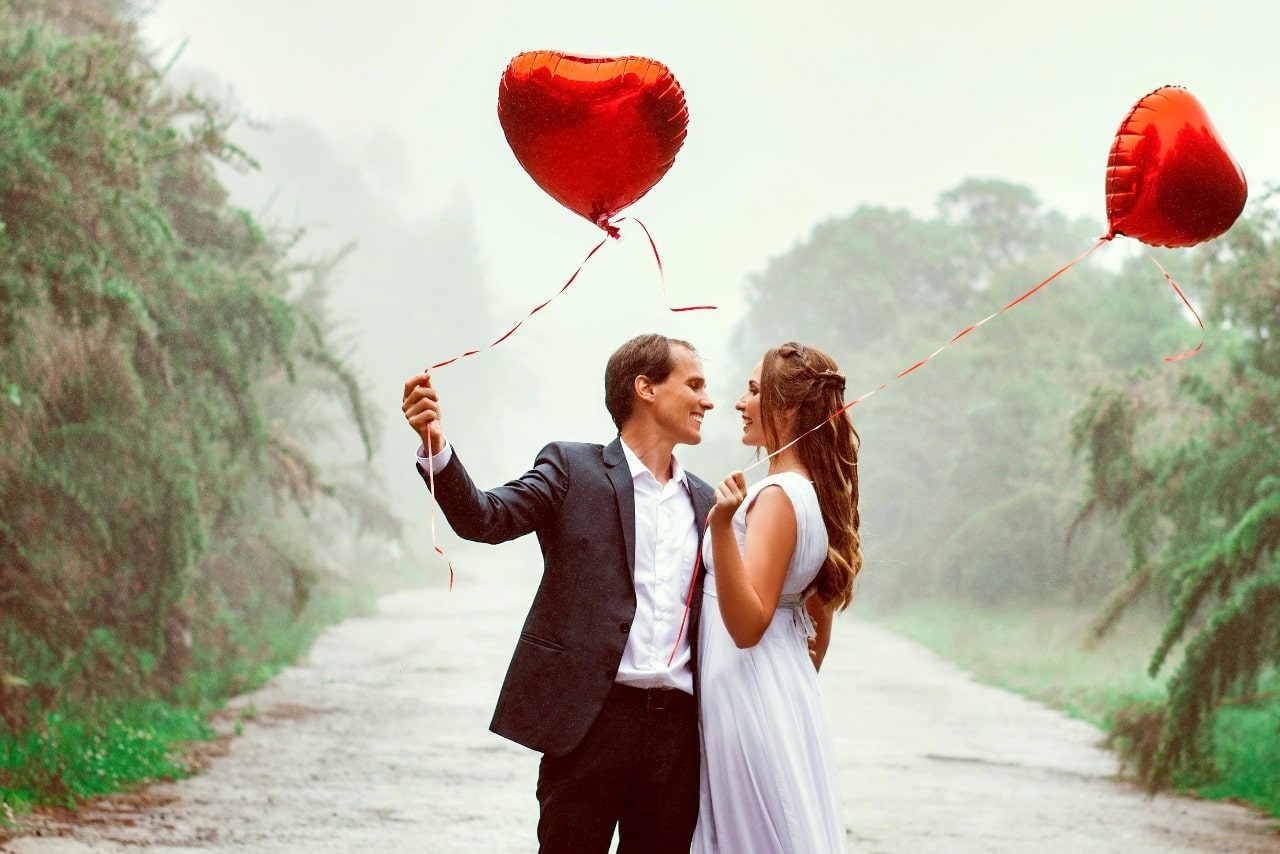 A newly engaged couple standing among dense shrubs, both holding red heart balloons.