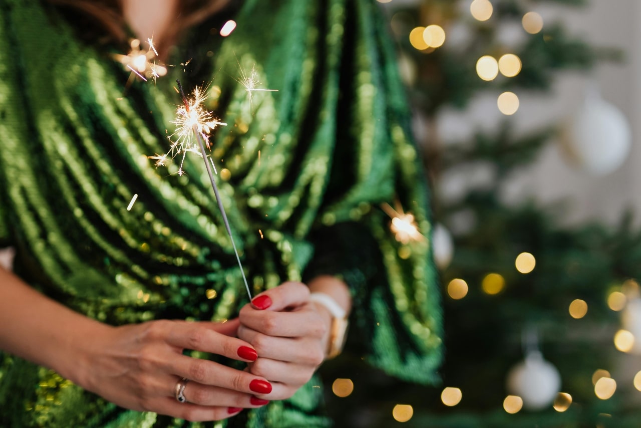 A close-up of a festively dressed woman in a green dress holding a sparkler in front of a Christmas tree.