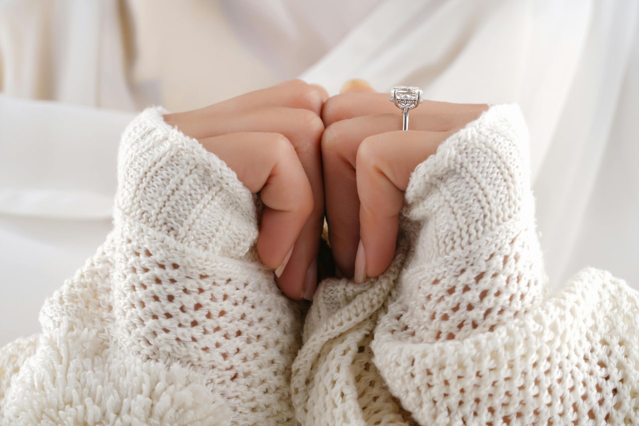 A close-up of a woman in a comfy sweater, wearing an elegant engagement ring on her right hand.