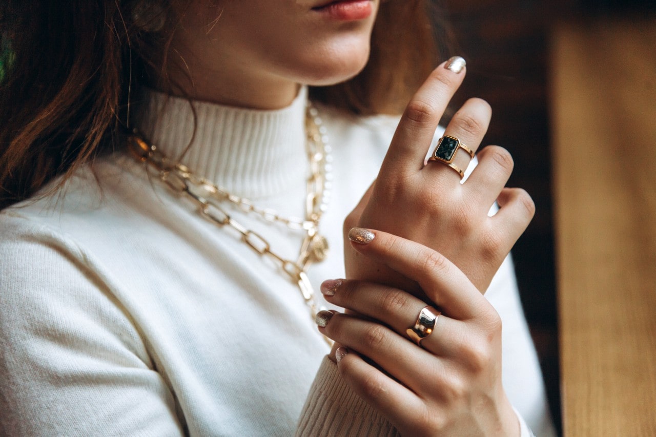 a woman looking out a window wearing chunky gold jewelry and a cream turtleneck