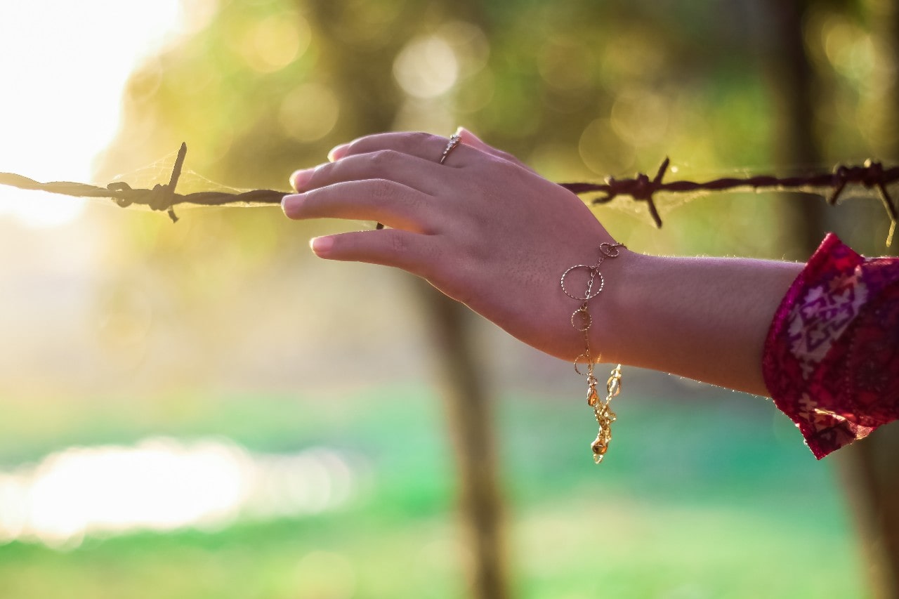 A woman wearing a yellow gold dainty bracelet and a yellow gold ring reaches out towards some barbed wire.
