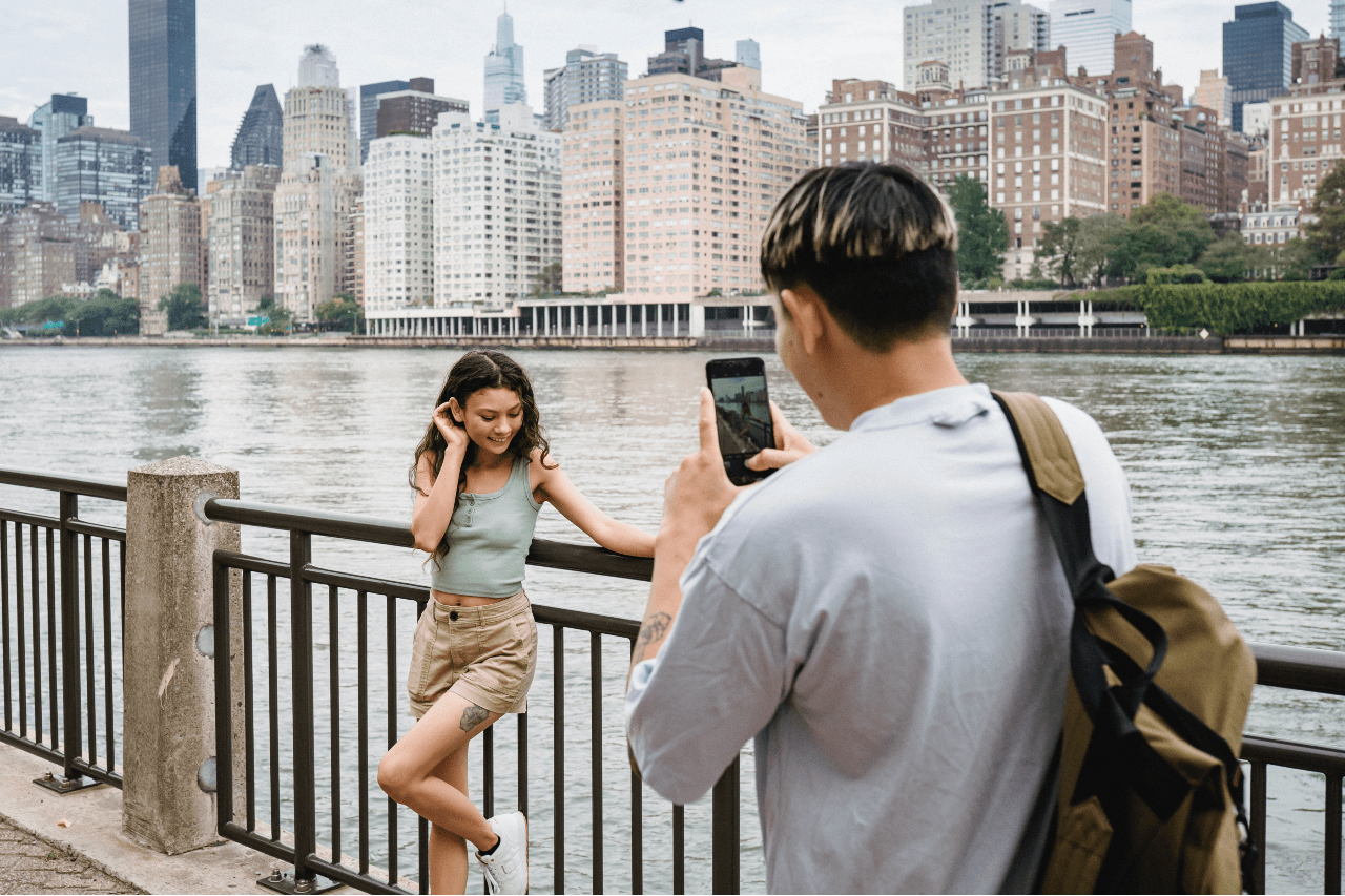 A woman poses while a man takes her picture next to a river by a big city.