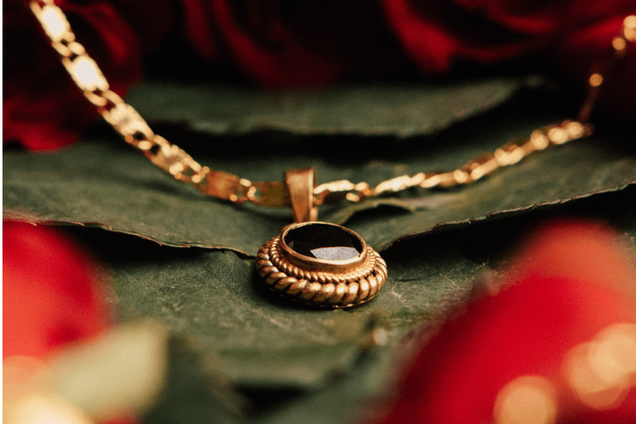 A close-up of an elegant gold necklace with a black gemstone pendant resting on green leaves surrounded by red rose petals.