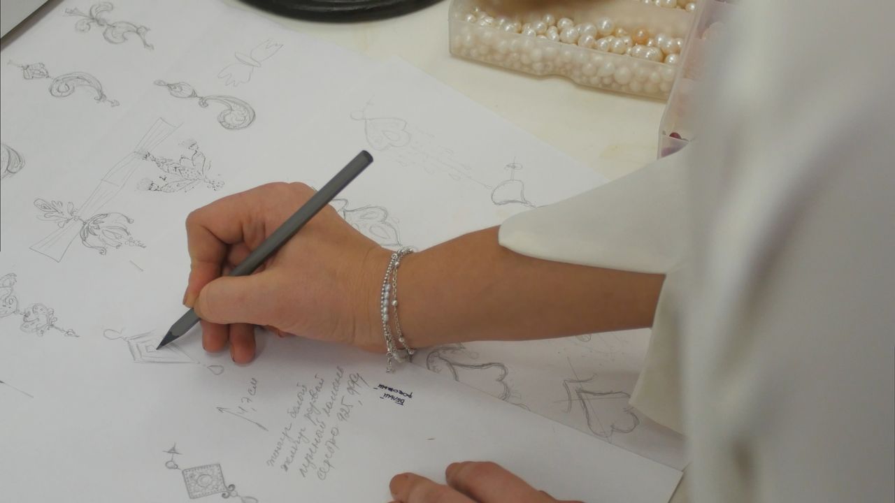 Hands sketch and annotate jewelry designs on paper, surrounded by box of pearls.