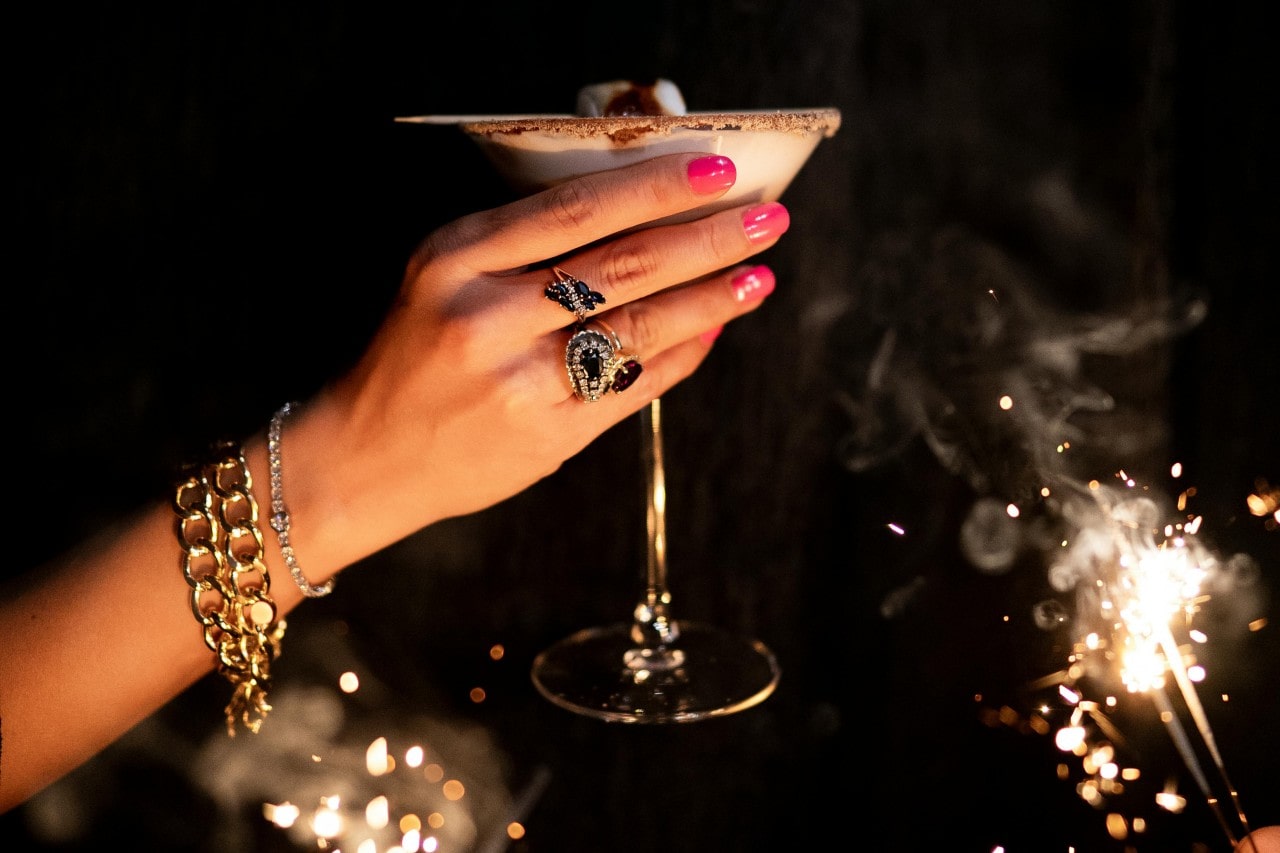 A woman holding a smores martini for a toast while wearing gold chain bracelets, a diamond tennis bracelet, and statement sapphire and diamond rings