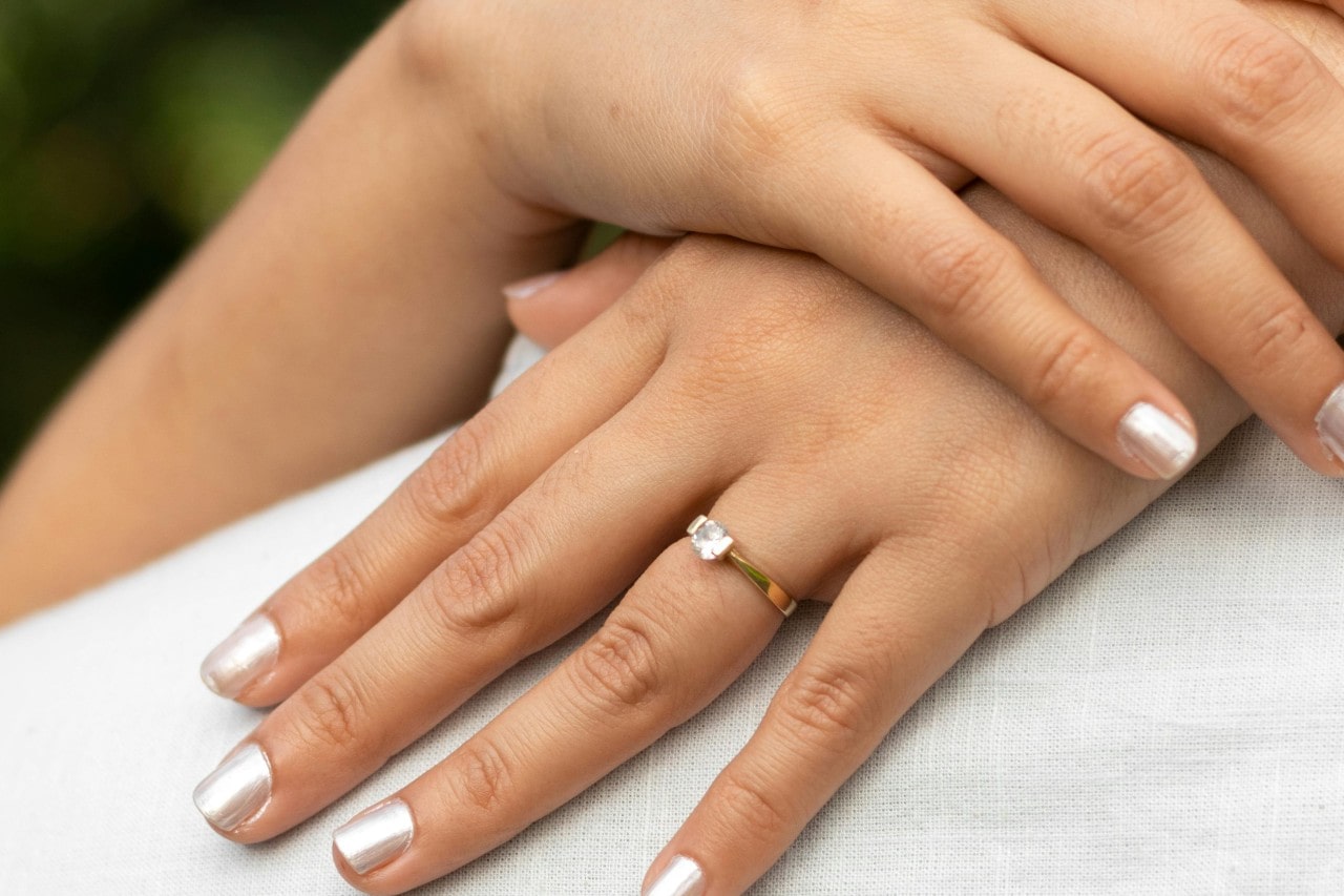 a woman’s hands around a man’s neck, wearing a yellow gold solitaire engagement ring