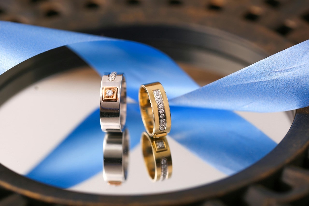 Two wedding bands with diamond accents on a mirror near a blue ribbon. Two wedding bands with diamond accents on a mirror near a blue ribbon.