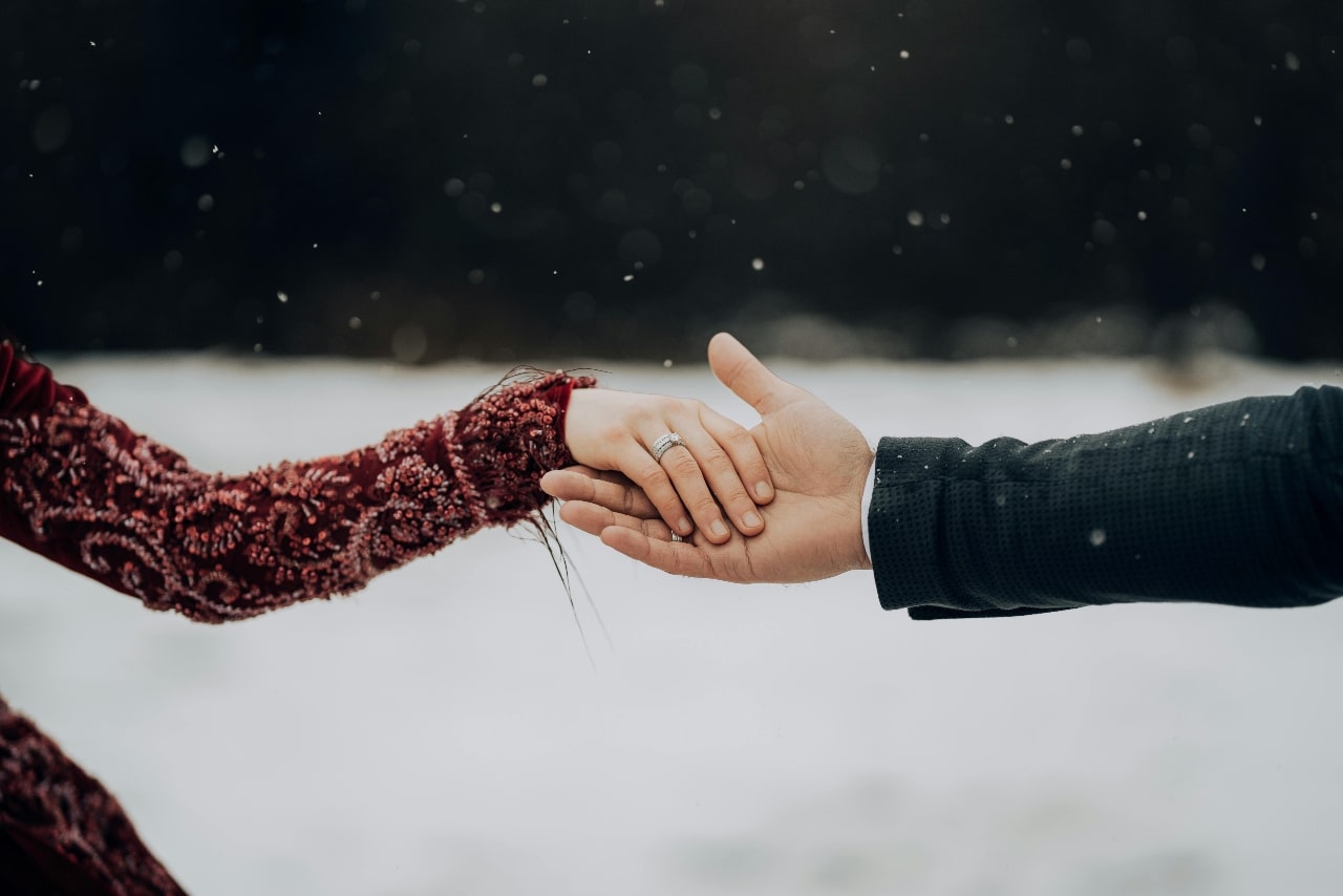 A couple holding hands in front of a snowy background, showcasing a white gold wedding band on a woman’s hand. A couple holding hands in front of a snowy background, showcasing a white gold wedding band on a woman’s hand.